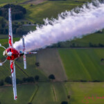 Red aerobatic plane trailing smoke over fields