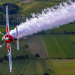 Red aerobatic plane trailing smoke over fields
